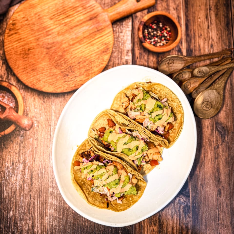 Three Señor Tomas Mexican tacos with guacamole and sauce on a white plate, on a wooden table.